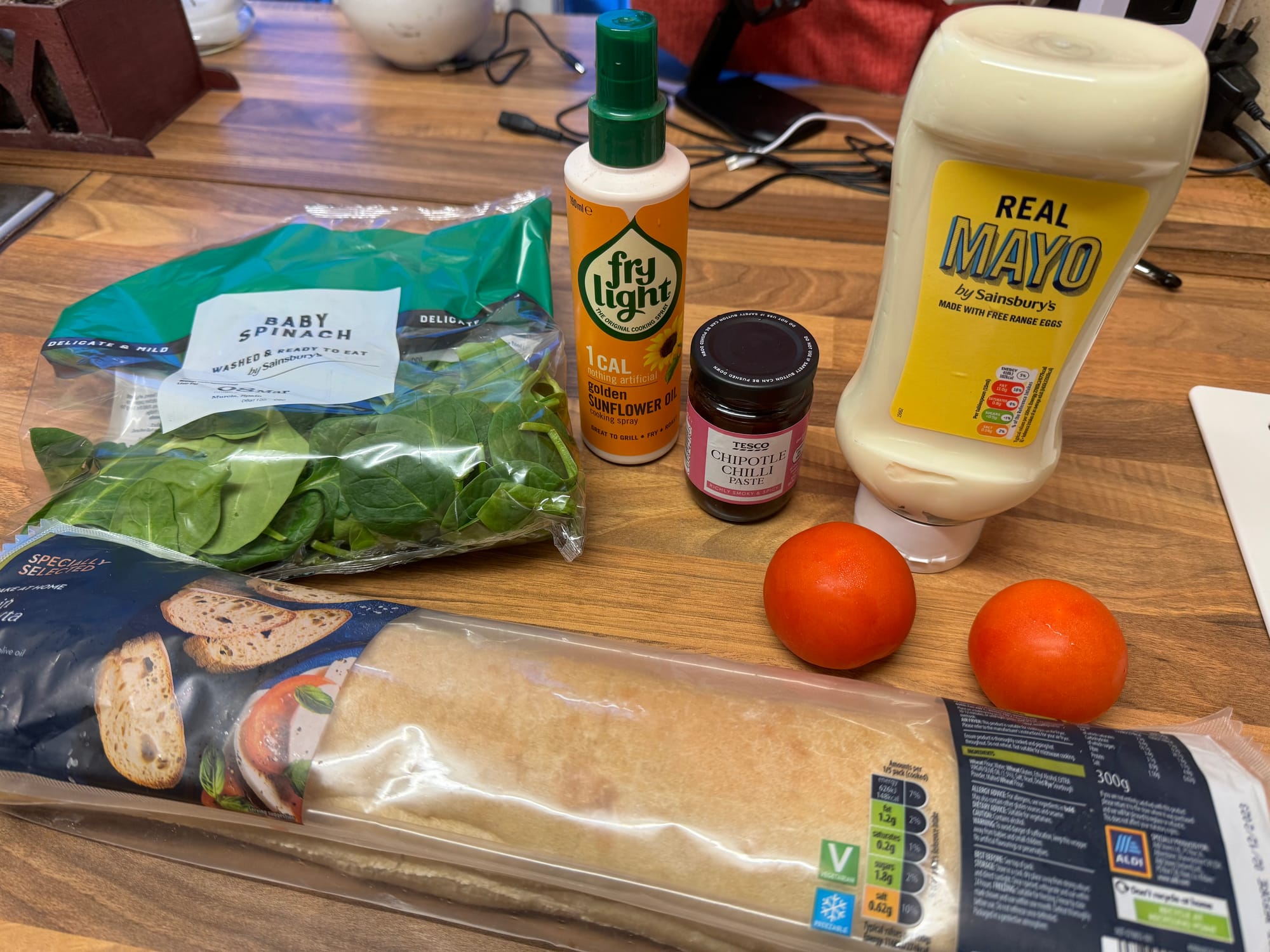 Ingredients for a sandwich on a table: spinach, tomato, bread, mayo, mustard, knife, cutting board.