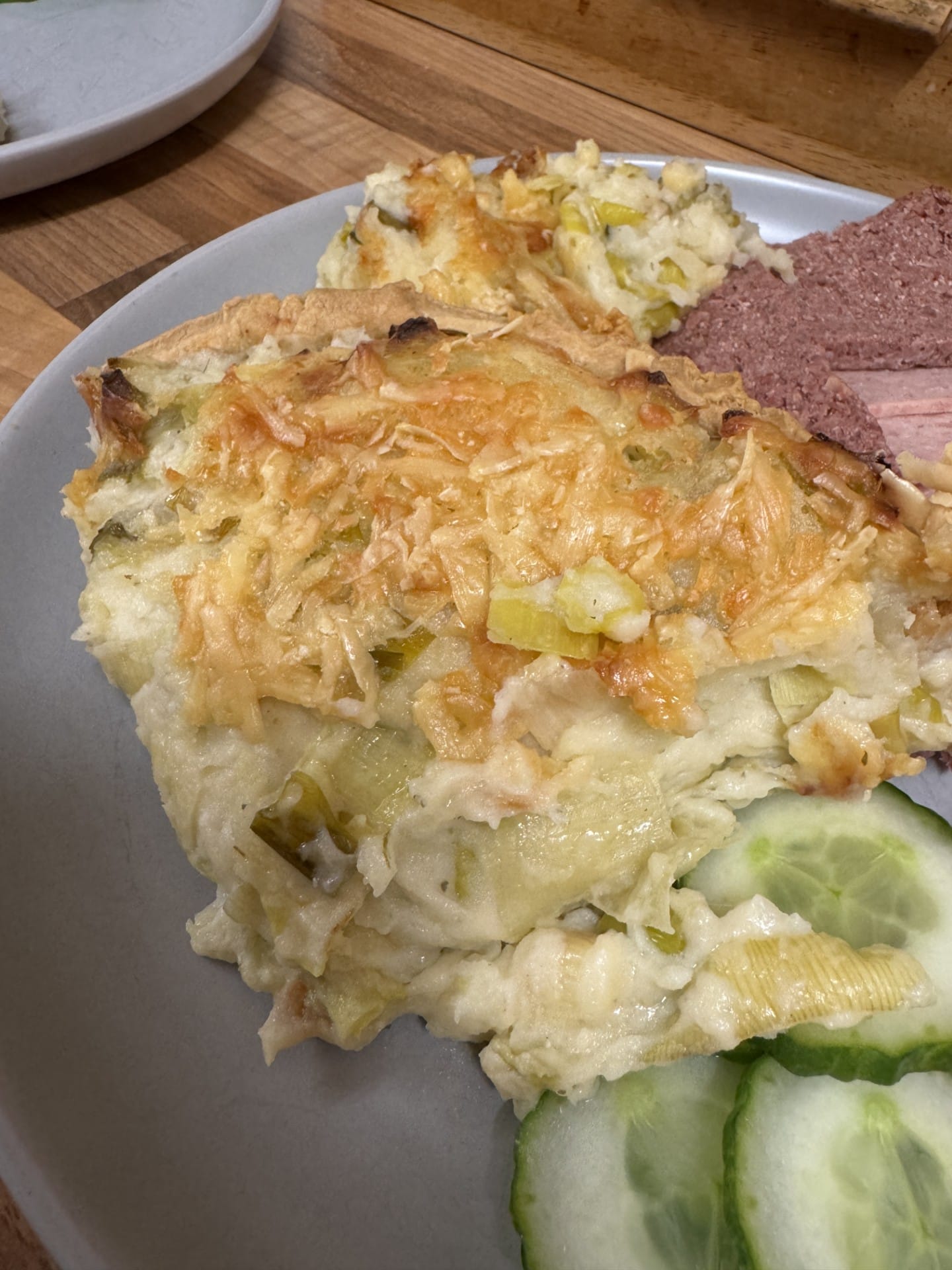 A close-up shot of a creamy slice of Potato, Leek and Two Cheese Pie showing the tender potato layers and a golden, bubbly melted cheese crust. Served on a rustic plate with a side of crisp cucumber and cured meat.
